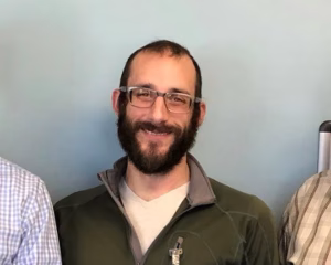 A bearded man wearing glasses smiles for a photograph against a plain background.
