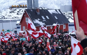 Crowd of protesters in Greenland holding red and white Greenlandic flags outside a snow-covered building, demonstrating against external pressure over Greenland&rsquo;s future.