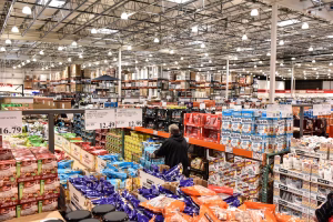 Interior of a Costco warehouse with aisles of bulk groceries, snacks, and household goods, with a shopper pushing a cart.