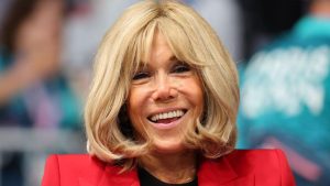 Brigitte Macron smiling in a red blazer during a public appearance.