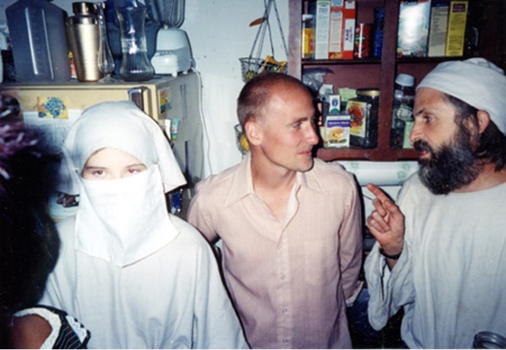 A veiled young person stands beside two adult men inside a small, cluttered kitchen, with shelves of food visible in the background.