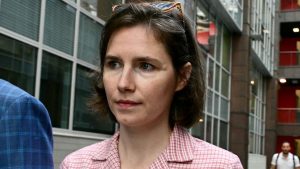 Amanda Knox walking outdoors in an urban setting, wearing a pink checkered shirt, photographed in daylight.