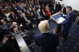A crowded press briefing room with journalists seated and photographing a speaker at the podium.