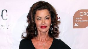 Janice Dickinson poses at a media event, wearing a black dress and red lipstick, photographed against a branded backdrop.