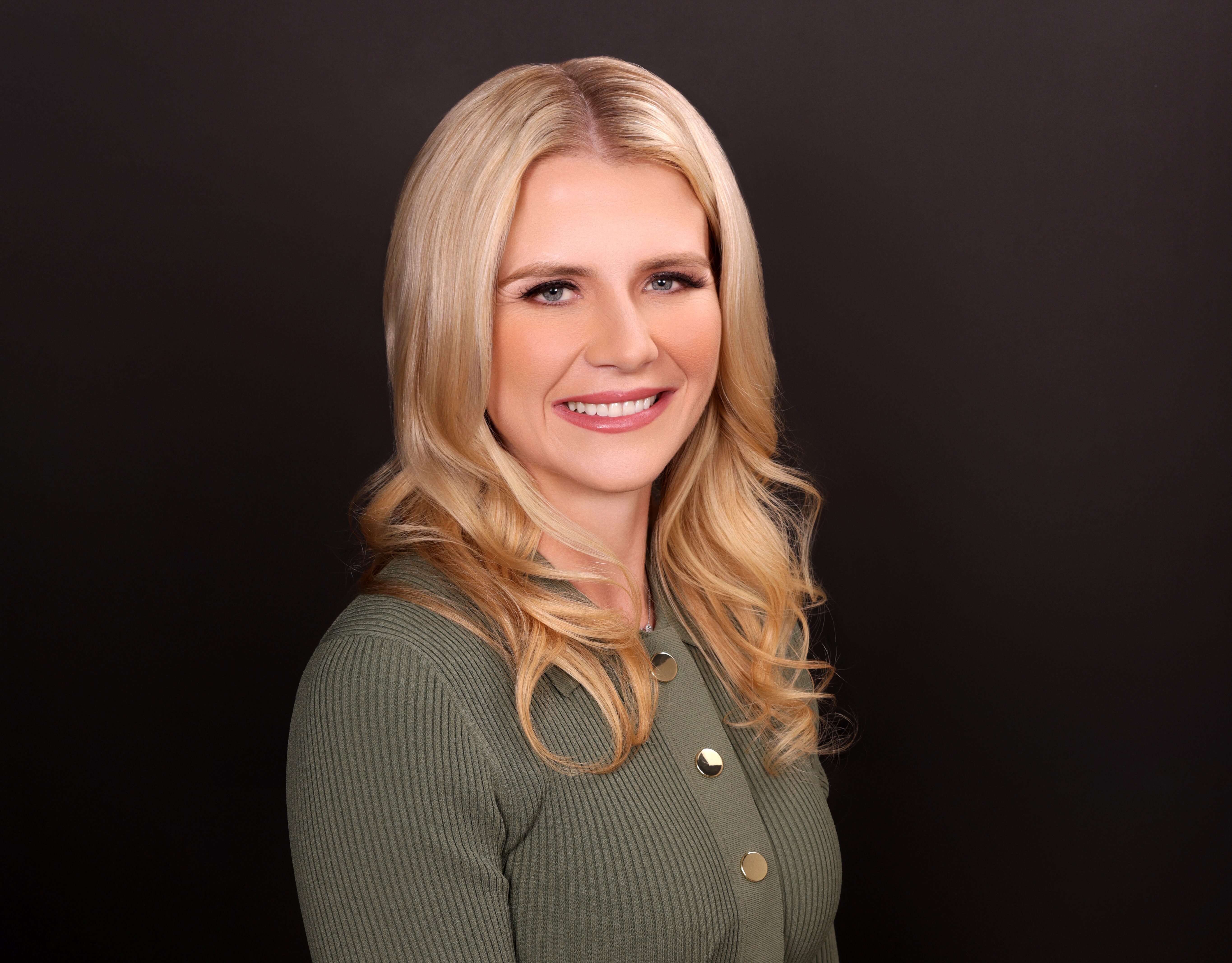 A professional portrait of a blonde woman smiling against a dark background, wearing a green top, photographed for advocacy or speaking materials.