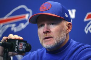 NFL head coach speaks at a press conference, holding a microphone against a Buffalo Bills backdrop.