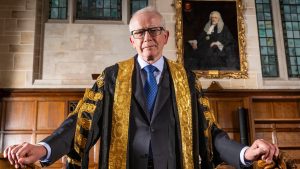 Senior UK judge wearing ceremonial robes standing in a historic courtroom with judicial portraits behind him