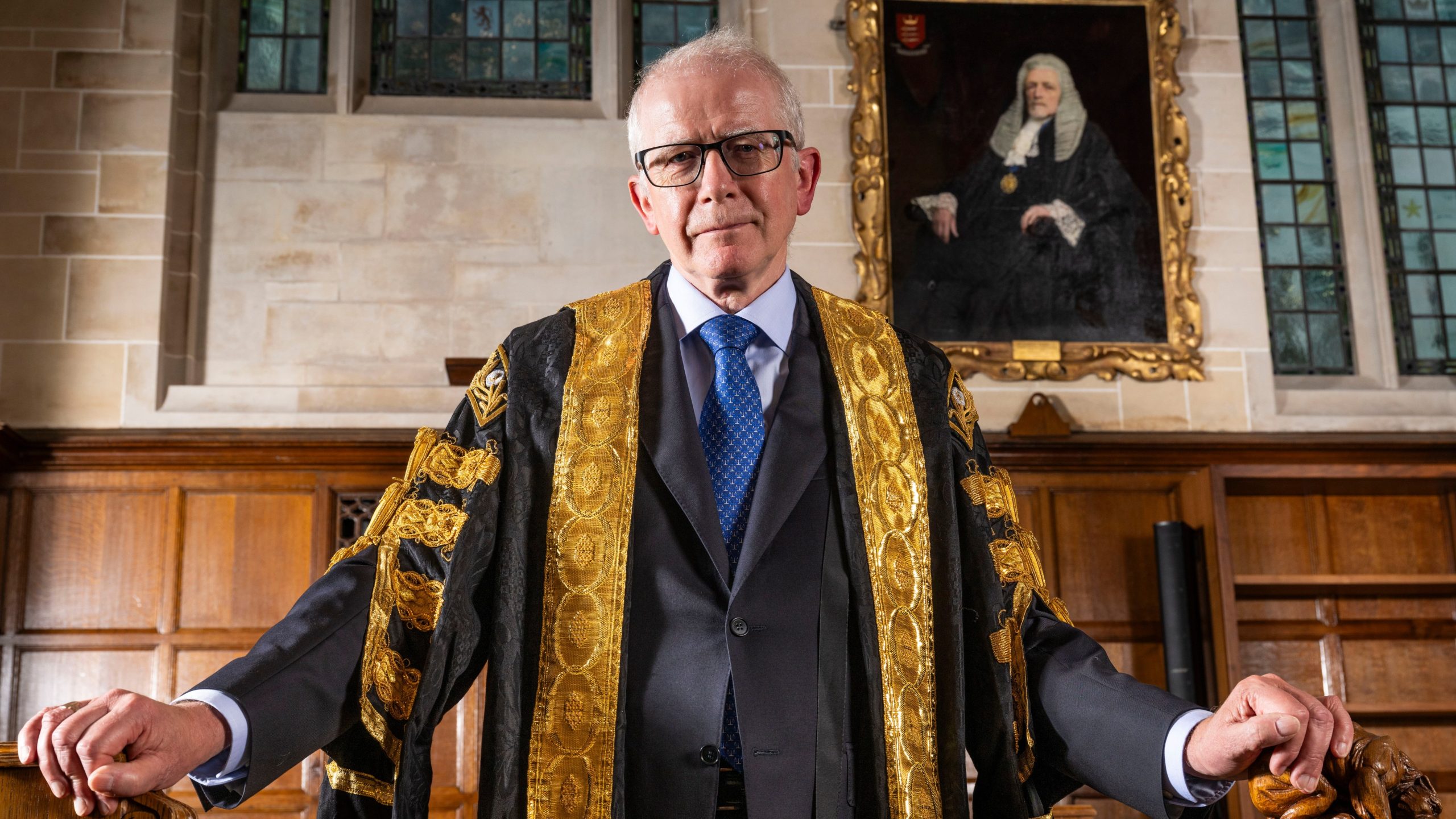 Senior UK judge wearing ceremonial robes standing in a historic courtroom with judicial portraits behind him