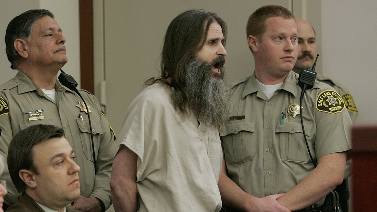 A bearded man in a jail uniform stands in a courtroom surrounded by sheriff’s deputies during a court appearance.