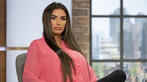 Katie Price appears during a studio interview, wearing a pink blouse and seated indoors with a city backdrop visible through a window.