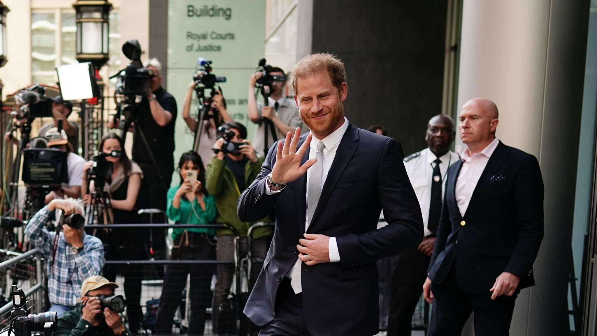 Man in a dark suit waves as he exits the Royal Courts of Justice in London, with photographers and security visible behind him.