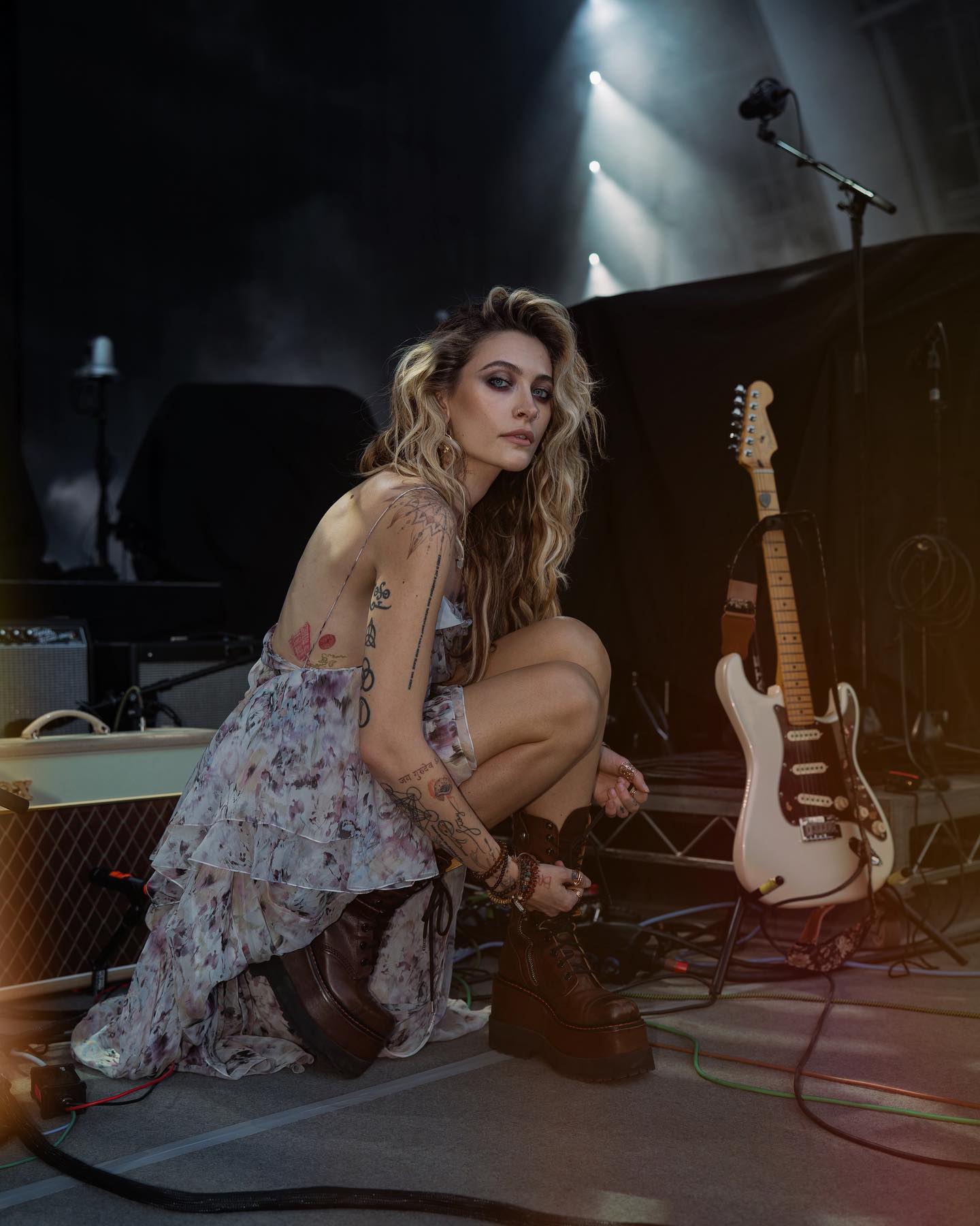 Paris Jackson performing on stage, wearing a floral dress and lace-up boots beside her guitar.