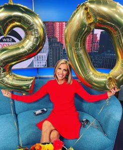 Kate Merrill seated on a studio couch holding large gold number balloons and smiling.