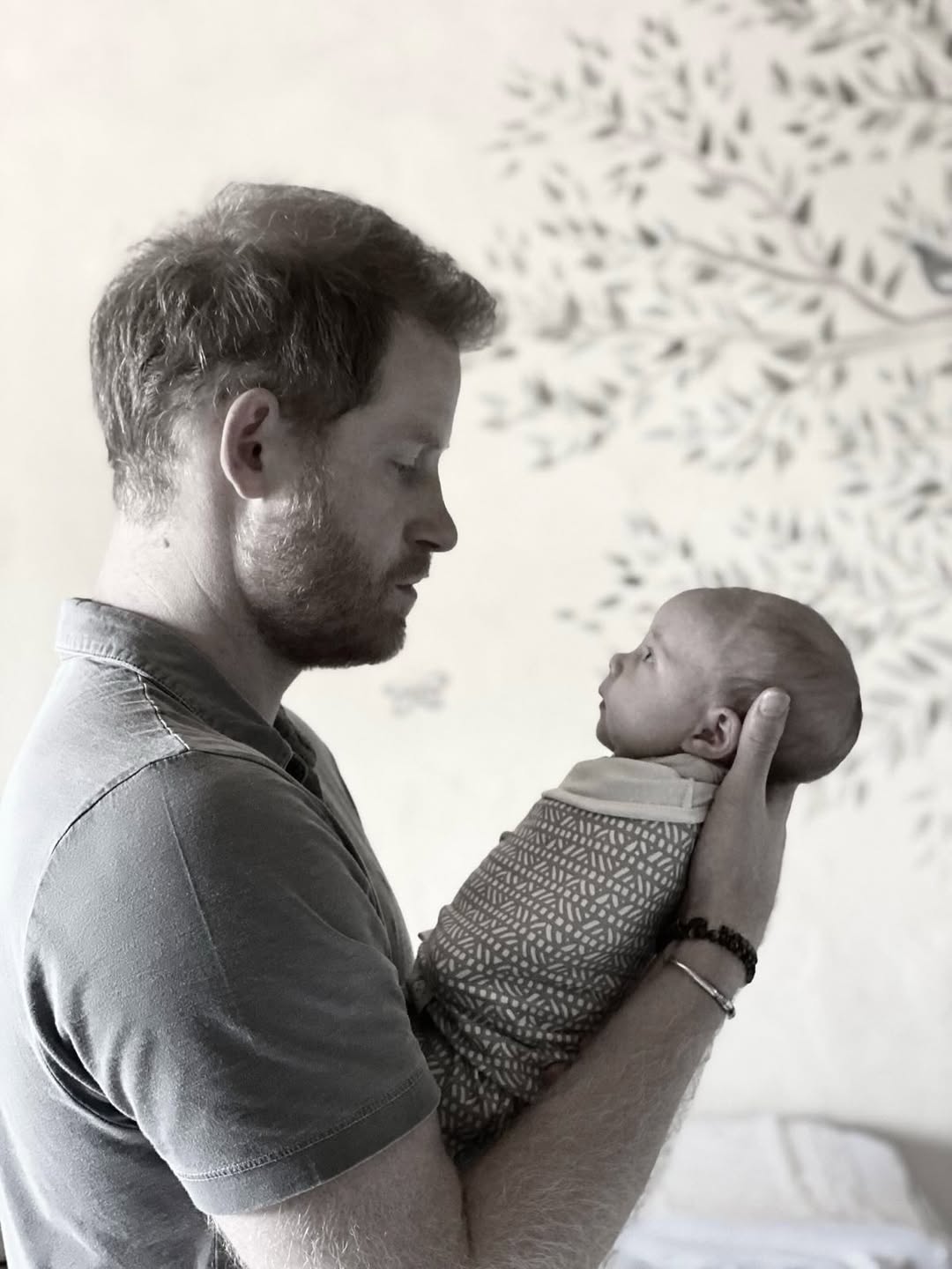 Prince Harry holding a newborn baby in a softly lit room with a tree mural in the background.