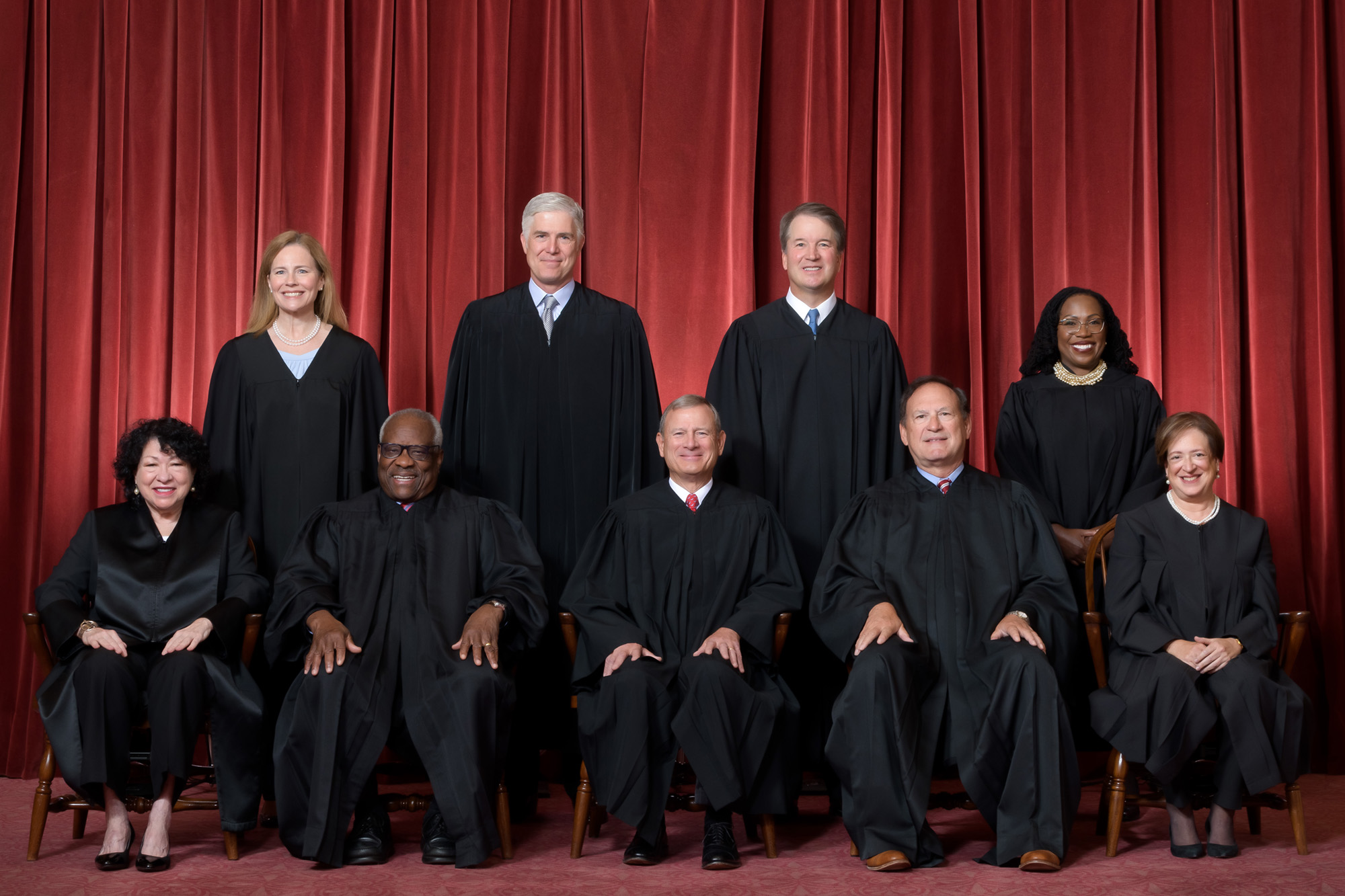 Group portrait of the U.S. Supreme Court justices seated and standing in judicial robes against a red curtain backdrop.