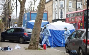 Forensic officers in protective suits walk past a cordoned street scene in London during a homicide investigation.