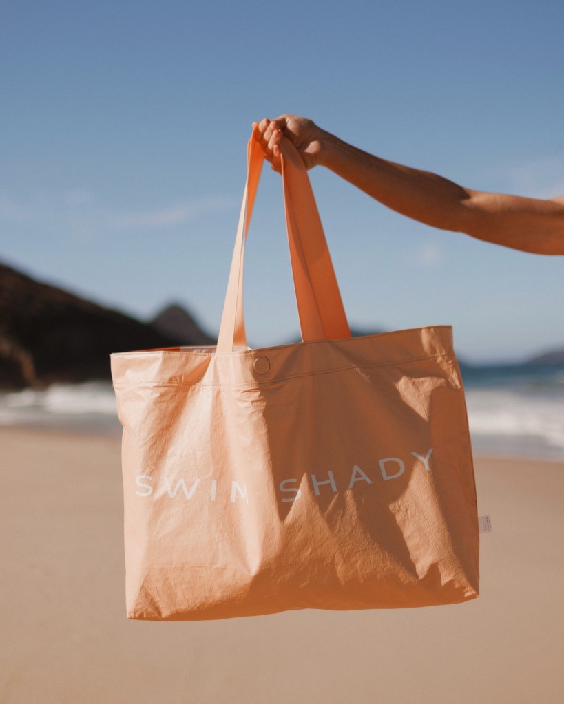 Person holding an orange Swim Shady beach tote bag on a sunny beach.